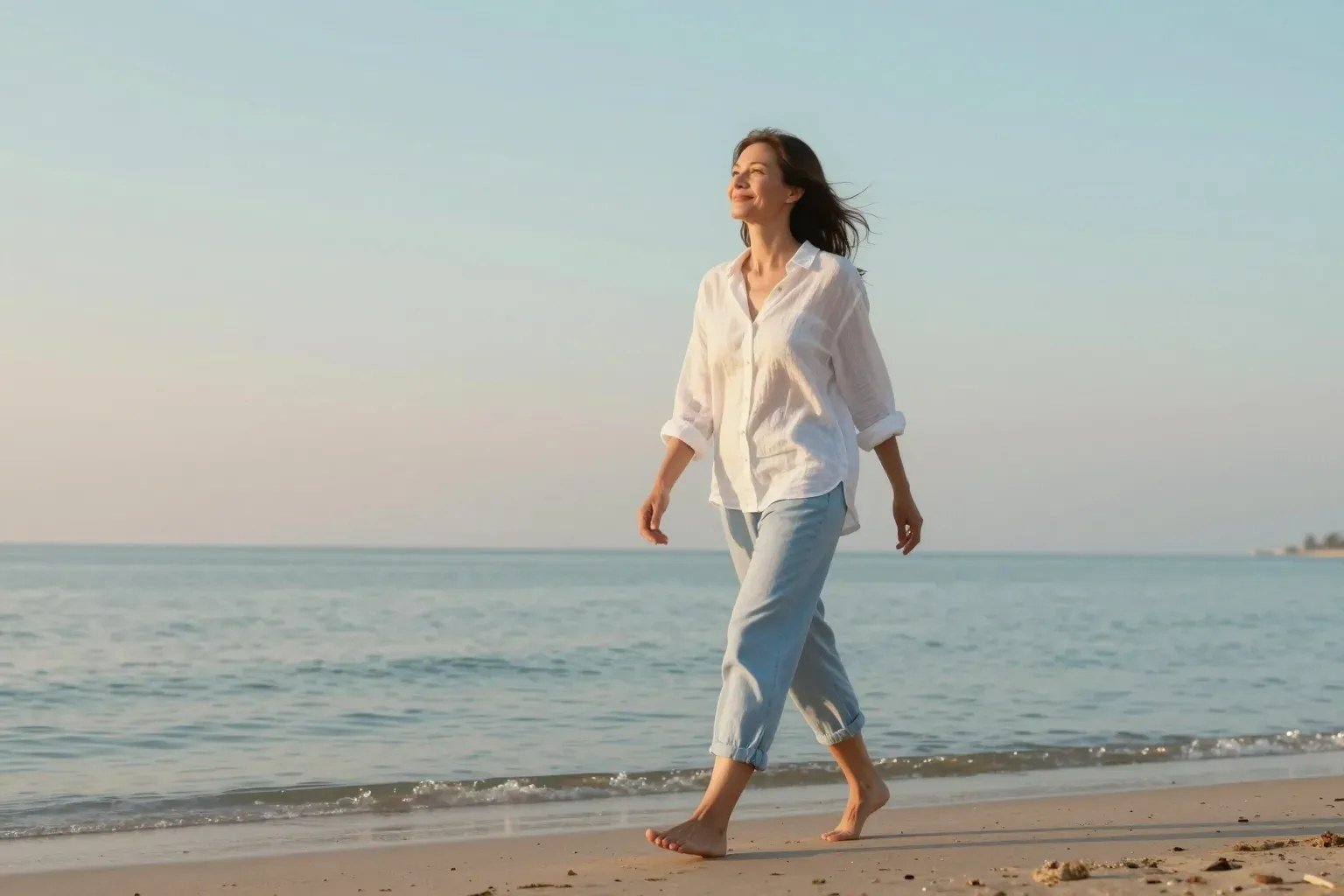 A woman walks barefoot along the shore in soft morning light, relaxed and smiling