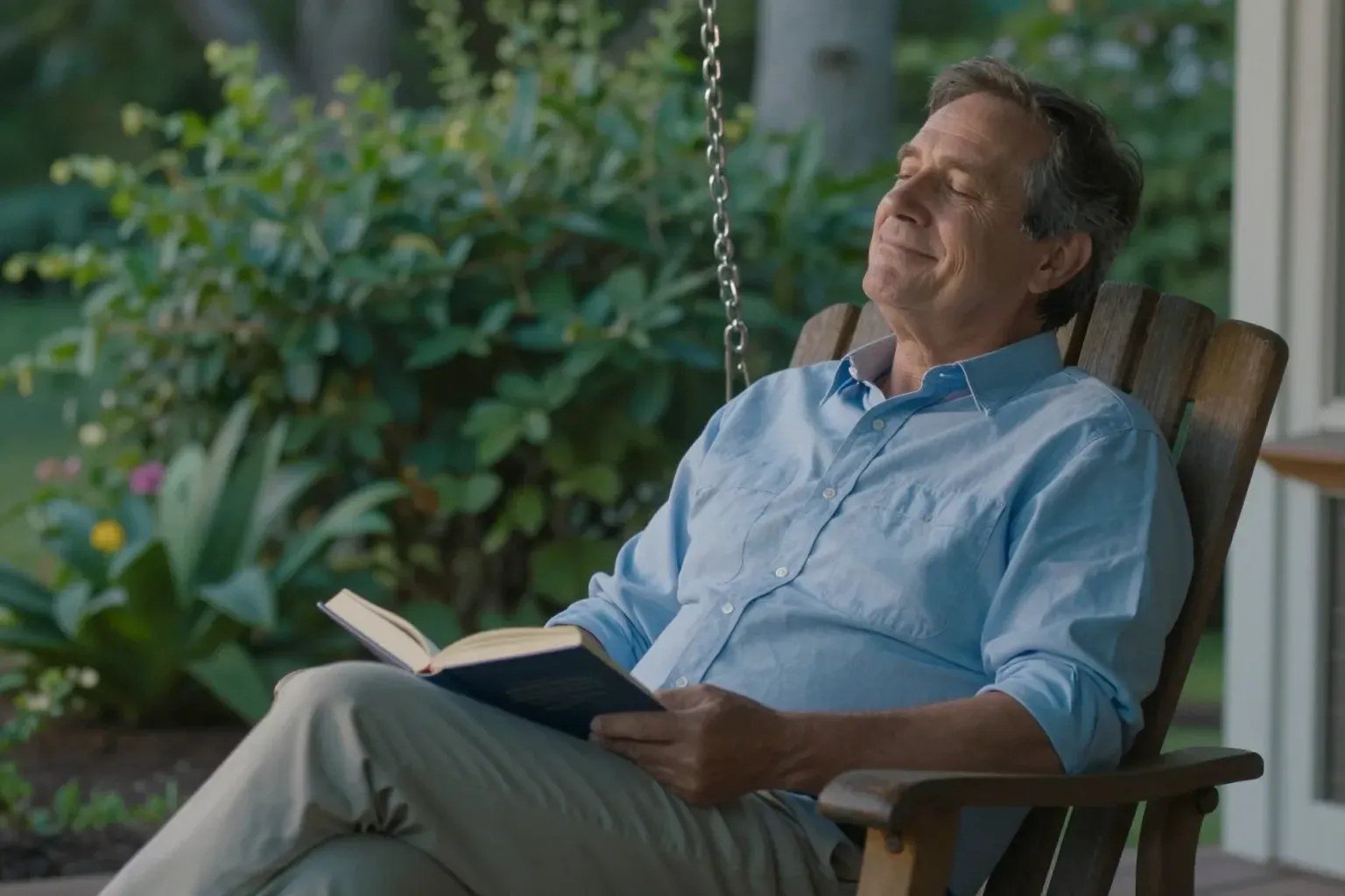 A man rests on a porch swing with a book in his lap, eyes closed and smiling in the garden air